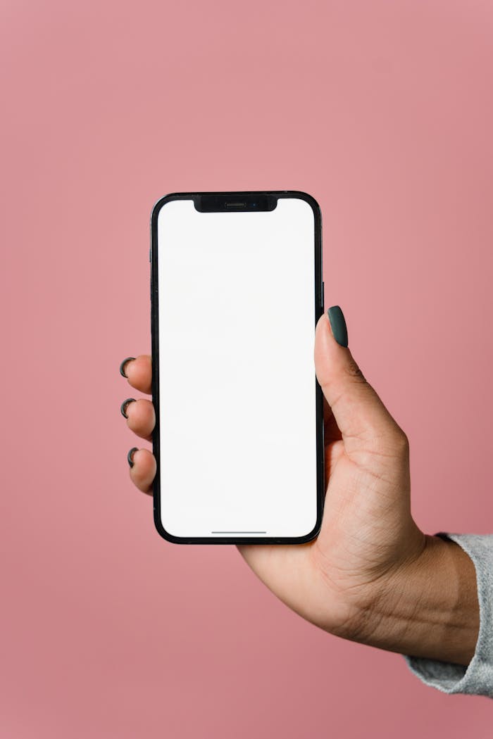 A hand holding a smartphone with a blank screen against a pink background.